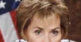 Judge Judy in a black robe with a lace collar, serious expression, sitting in court with an American flag backdrop.