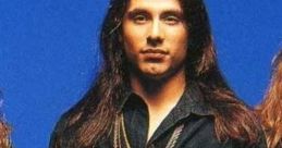 Andre Matos of Shaman poses confidently, showcasing his signature long hair and distinct black outfit against a blue backdrop.