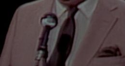 Edward Herlihy narrating a newsreel in 1965, wearing a suit and smiling, speaking into a microphone on stage.