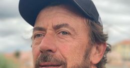 Benny Benassi with a beard, wearing a black cap and shirt, gazes thoughtfully outdoors against a cloudy sky backdrop.