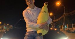 Young man in a hoodie poses with a plush banana on a car, showcasing casual nighttime style and urban vibes.