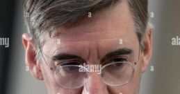 Jacob Rees-Mogg in a formal setting, wearing glasses, a striped shirt, and a tie, looking thoughtful and engaged.