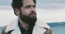 Sol Rosenberg outdoors, wearing a stylish brown jacket with a shearling collar, gazing thoughtfully at the scenic background.