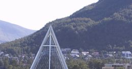 Modern glass pyramid building in Norway, surrounded by trees and buildings, with mountains in the backdrop.