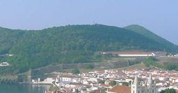 Scenic view of AF61 village, showcasing white buildings, red roofs, and lush green hills in the background.