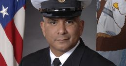 Portrait of a uniformed Navy officer with medals, against a backdrop featuring the American flag and an emblematic banner.