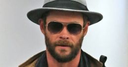 Stylishly dressed man with a beard in a gray hat and sunglasses, showcasing a modern, urban fashion aesthetic.