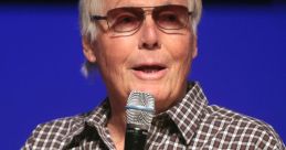 Adam West speaking at an event, wearing sunglasses and a plaid shirt, holding a microphone, showcasing his iconic charm.