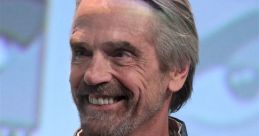 Jeremy Irons smiles on stage, wearing a stylish blue outfit and necklace, exuding charm and charisma during a public event.