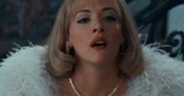 Debbie Jellinsky in glamorous white attire with a feathered wrap and pearl necklace, exuding elegance and allure.