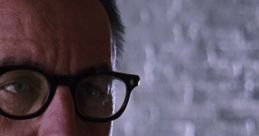 Warden from Shawshank Redemption, wearing glasses, with a serious expression in a stark, gray setting.