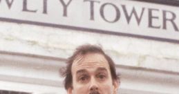 Basil Fawlty stands in front of Fawlty Towers, showcasing his iconic comedic expression and classic striped tie.