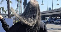 Long-haired individual wearing black, walking down a sunny street with palm trees and a bag, embodying a relaxed vibe.