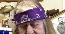 Man with long beard and purple bandana, seated in a cluttered room, reflecting a unique and unconventional style.