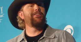 Toby Keith, country music star, speaks at a press event wearing a cowboy hat and denim jacket. Engaging personality shines through.