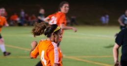 Female soccer player in an orange jersey dribbles the ball on a green field during a night match, showcasing football skills.