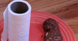 Chocolate treat shaped like poop on a red plate, next to a roll of paper towels, playful and humorous food presentation.