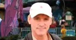 Chris The Hacker smiling at a vibrant amusement park, wearing a cap and holding a drink, surrounded by colorful attractions.