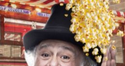 Danny DeVito playfully holds his hand up as popcorn spills around him, dressed in a vibrant red coat and hat, at a fair.