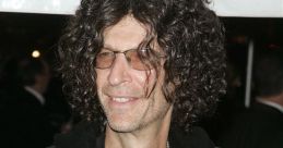 Howard Stern smiling at an event, wearing glasses and a black jacket with curly hair, exuding his signature style.