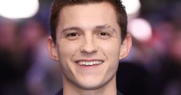 Tom Holland smiles warmly at an event, wearing a navy blazer over a grey shirt, radiating charm and confidence.