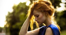 A woman in a blue dress stands in a sunlit park, thoughtful and serene, with flowing red hair and an elegant pose.