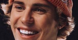 Justin Bieber smiles brightly, wearing an orange beanie and white sweater, showcasing his tattoos and youthful charm.