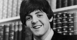 Paul McCartney smiling while playing a bass guitar, with bookshelves in the background, showcasing his musical talent.