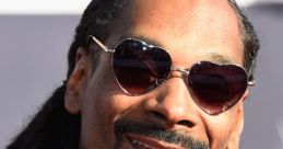 Snoop Dogg smiles confidently, showcasing his signature style with heart-shaped glasses and iconic dreadlocks at a red carpet event.