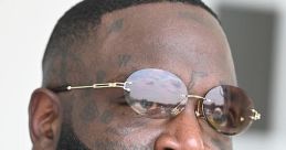 Rick Ross sporting stylish sunglasses and a bold beard, showcasing his signature look with intricate tattoos and chains.