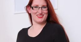 Woman with long red hair and glasses wearing a black top and metallic skirt, posing confidently in a bright room.