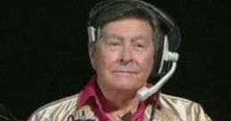 Johnny Gilbert wearing a gold jacket and headset, hosting a game show with a microphone and computer in front of him.