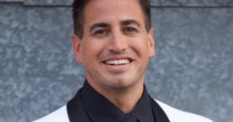 Justin Roberts smiles confidently in a stylish black tie and white tuxedo against a textured gray background.