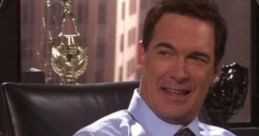 Patrick Warburton smiling in a blue shirt, seated in an office with trophies and a coffee cup in the background.