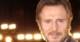 Liam Neeson posing confidently, wearing a stylish suit, illuminated by bright lights at a red carpet event.