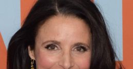 Julia Louis-Dreyfus smiles warmly, showcasing her elegant look with a black outfit and golden jewelry at a red carpet event.