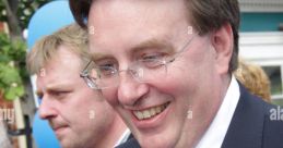 John Michael Howell smiling at an event, wearing a blue rosette, with a cheerful atmosphere and balloons in the background.