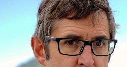 Louis Theroux with a serious expression, wearing glasses and a blue shirt, against a scenic mountainous background.