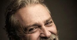 Haluk Bilginer smiles warmly, showcasing his distinct beard and charming personality in a close-up portrait.