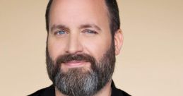 Tom Segura smiling, wearing a black jacket, showcasing his beard and blue eyes against a soft beige background.