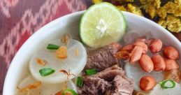 A bowl of soto with tender beef, radish, crispy shallots, green onions, and lime on a vibrant patterned tablecloth.