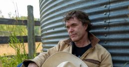 Cory Lunny casually rests by a weathered silo, holding his cowboy hat, surrounded by lush grass and scenic rural vibes.