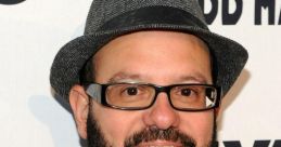 David Cross wearing a stylish hat and glasses, smiling at a promotional event backdrop with bold text.