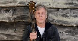 Paul McCartney holding an acoustic guitar, standing against a rustic wooden backdrop, showcasing his musical artistry.