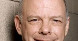 Wallace Shawn smiling, wearing a blue jacket, known for voicing Rex in Toy Story and his work in theater and film.