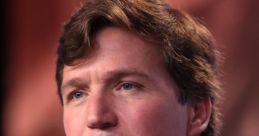 Tucker Carlson speaking into a microphone at a public event, wearing a suit and a tie, with a focused expression.