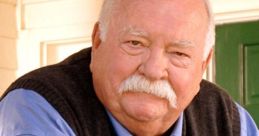 Wilford Brimley smiles warmly, wearing a blue shirt and gray vest, embodying his beloved, approachable personality.