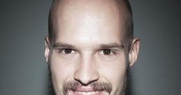 Portrait of Daniel Cz with a shaved head and beard, smiling in a smart black suit and white shirt against a dark background.