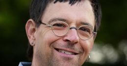 Portrait of Tobias Wredenmark smiling outdoors, wearing a blue patterned shirt, glasses, and an earring, with greenery in background.