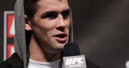 Dominick speaking at a UFC event, wearing a hoodie and a graphic t-shirt, holding a microphone, engaging the audience.